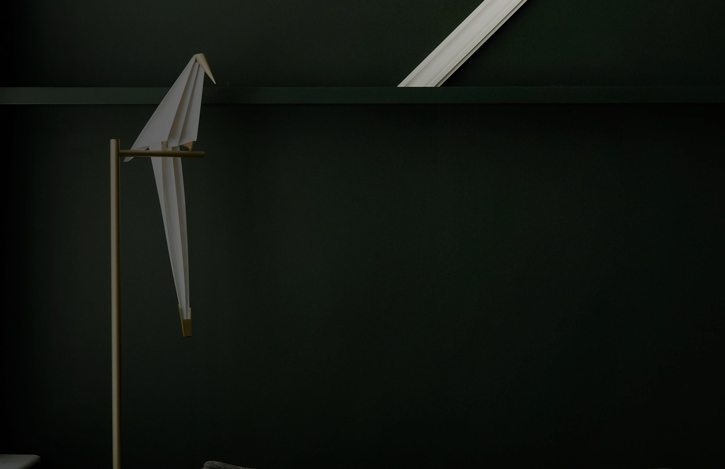 A white origami paper crane perched on a slim brass floor lamp against a dark green wall